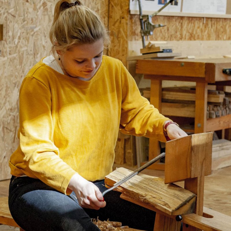 Eine Frau sitzt auf einer Werkbank uns schnitzt an einem Stück Holz